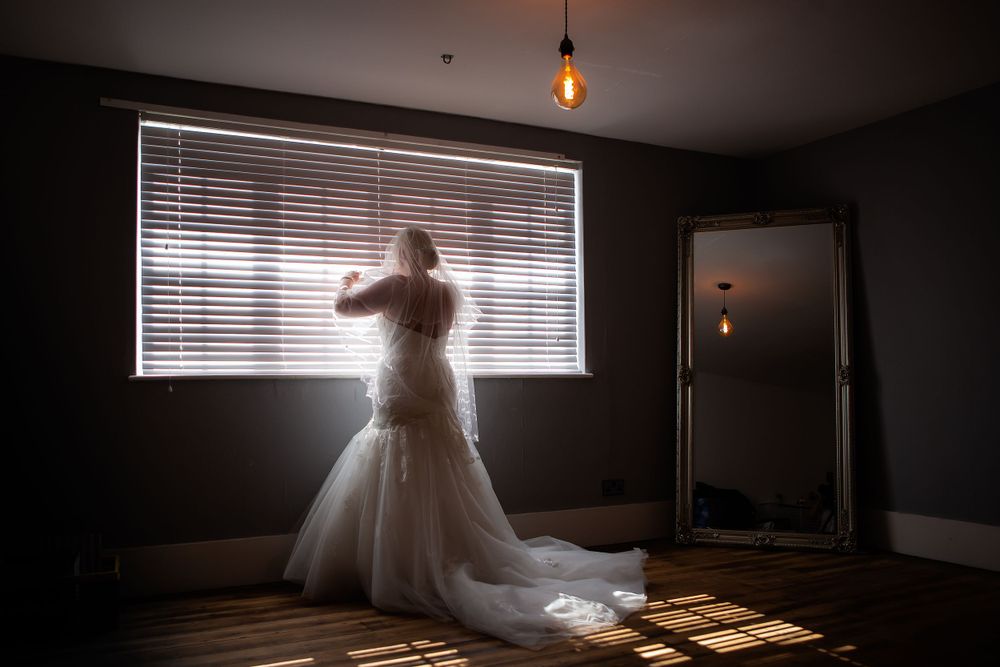 A silhouette of a bride in a wedding gown standing by a window with venetian blinds.
