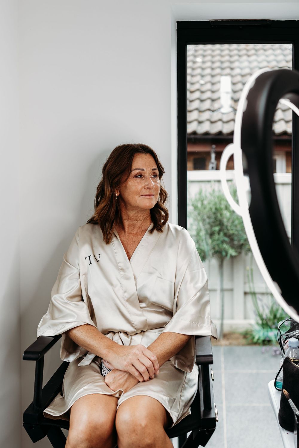 A woman in a cream-colored robe sits in a chair during wedding preparations.