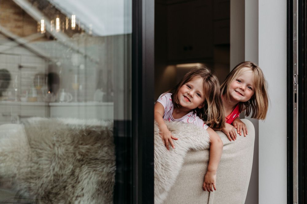 Two children wrapped in blankets peeking through a glass door with joyful expressions.