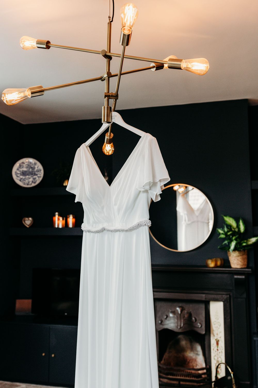 An elegant white wedding dress hangs from a modern brass light fixture against a dark wall.