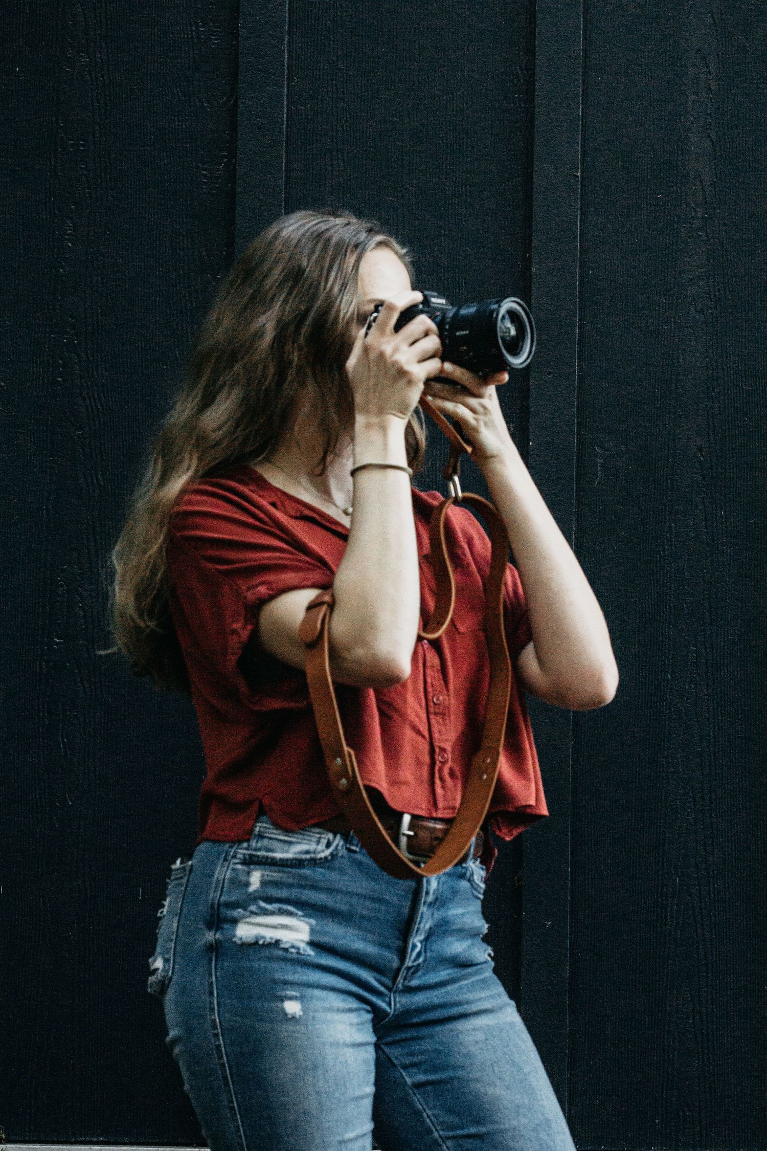 A photographer looks through her camera lens in front of a dark wall, moody aesthetic