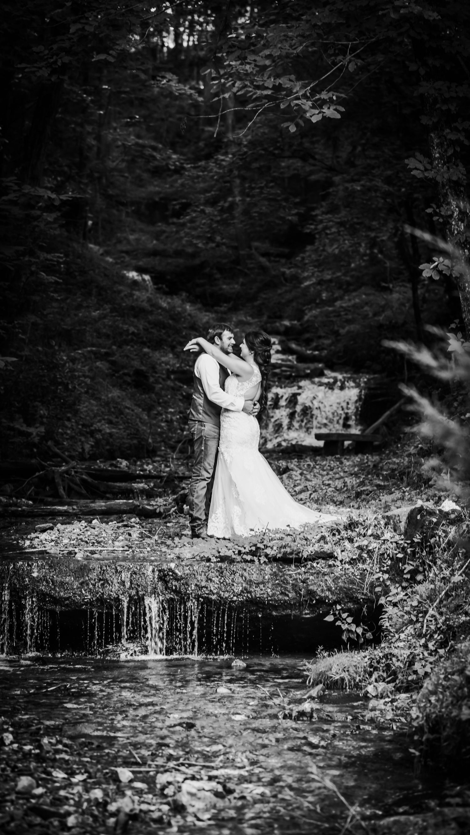 Black and white wedding portrait of couple by cascading waterfall in forest setting.