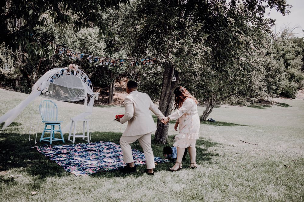 Man in tan suit dancing joyfully with woman in floral dress on a colorful blanket in a sunny garden.