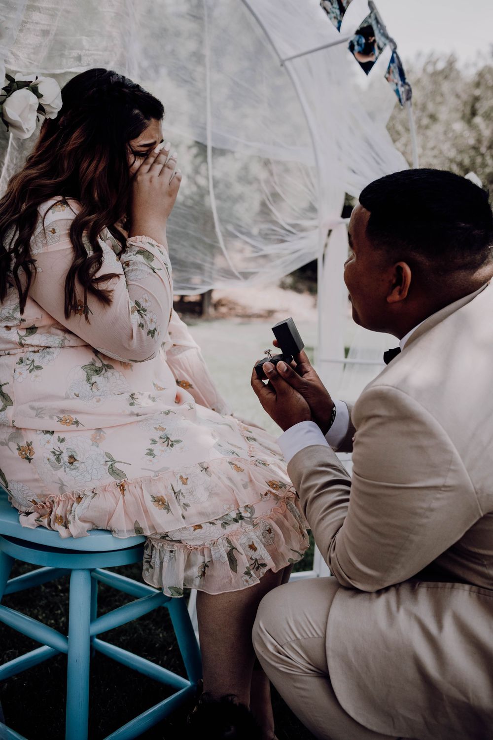 Man kneeling proposing with ring box to emotional woman in floral dress during romantic outdoor proposal.