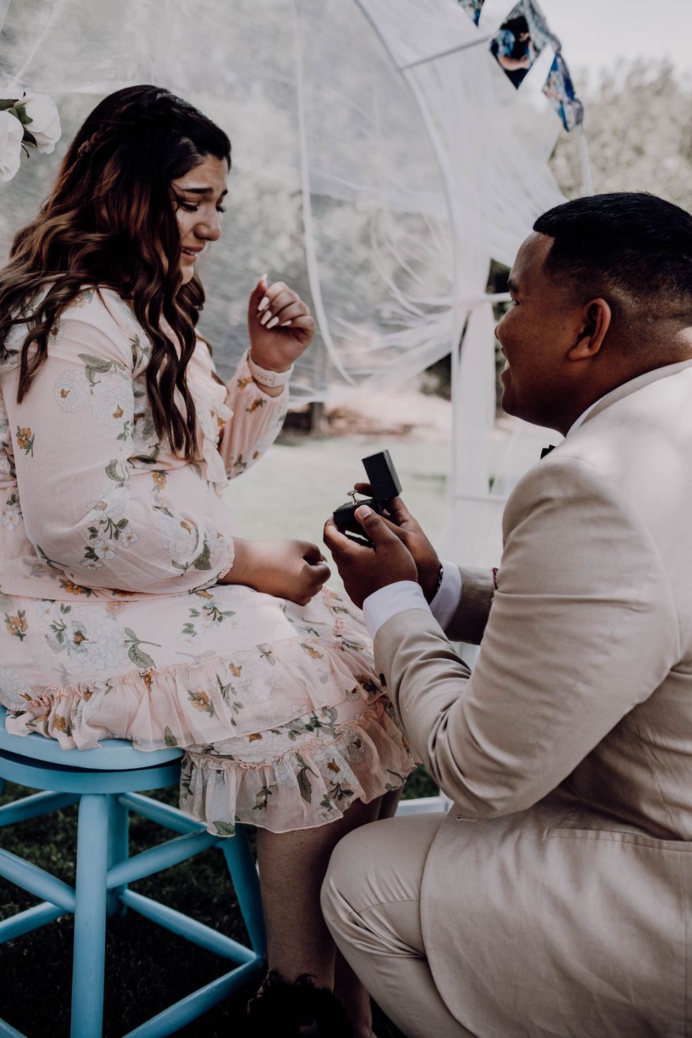 Man kneeling and presenting engagement ring box to emotional woman seated on blue stool during outdoor proposal.