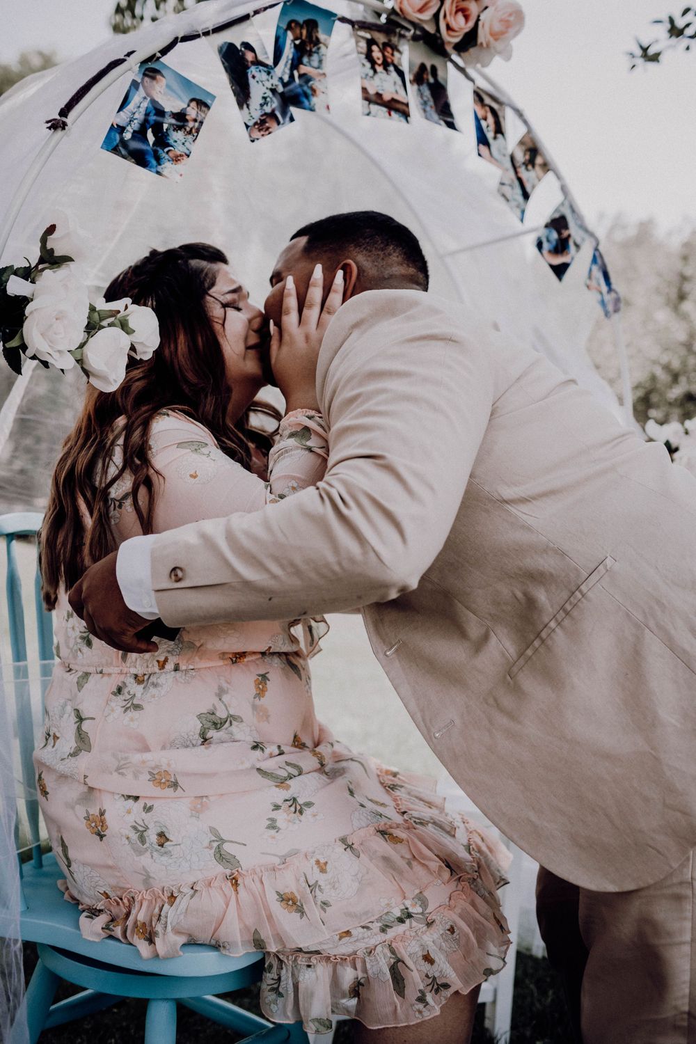 Couple sharing first kiss after engagement under decorated arch with photo garland outdoors.
