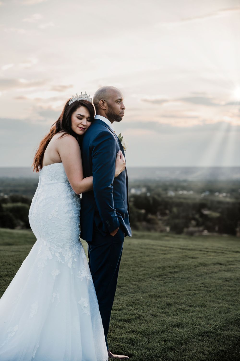 A wedding couple poses together on a grassy overlook during sunset.