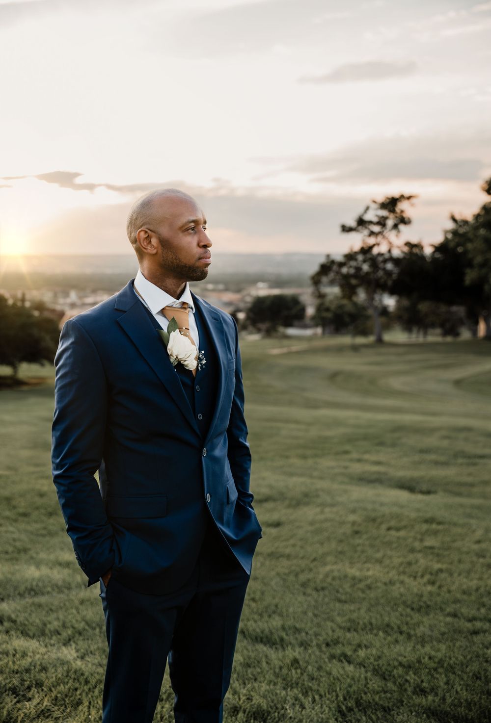Elegant groom in navy suit poses at sunset on hilltop overlooking scenic landscape.