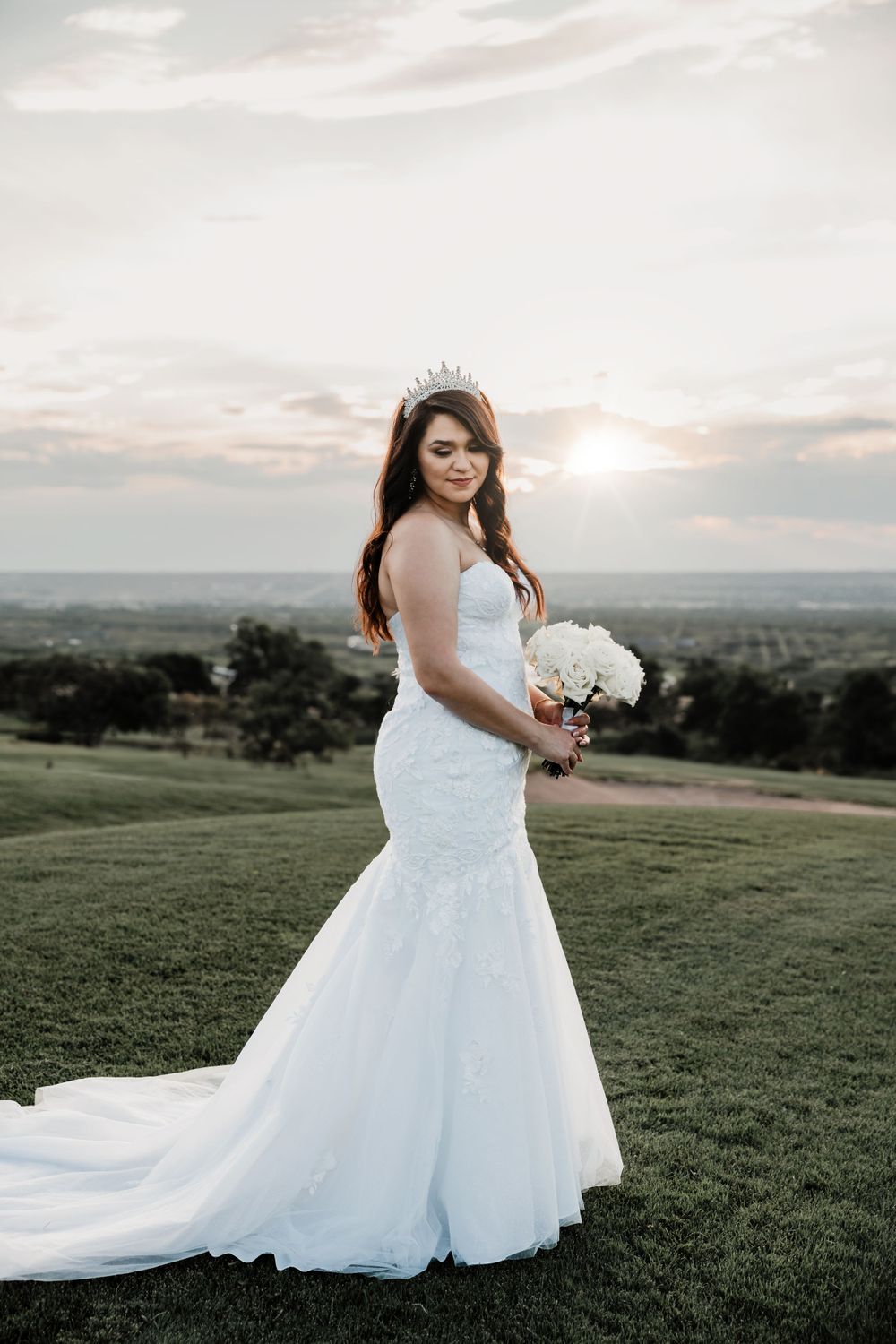 Elegant bride in fitted lace gown poses with bouquet during sunset on rolling hillside.