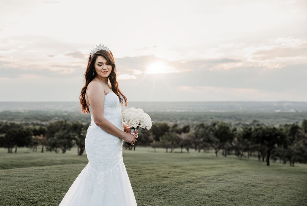 A bride in a white mermaid dress holds a bouquet while standing on a hilltop at sunset.