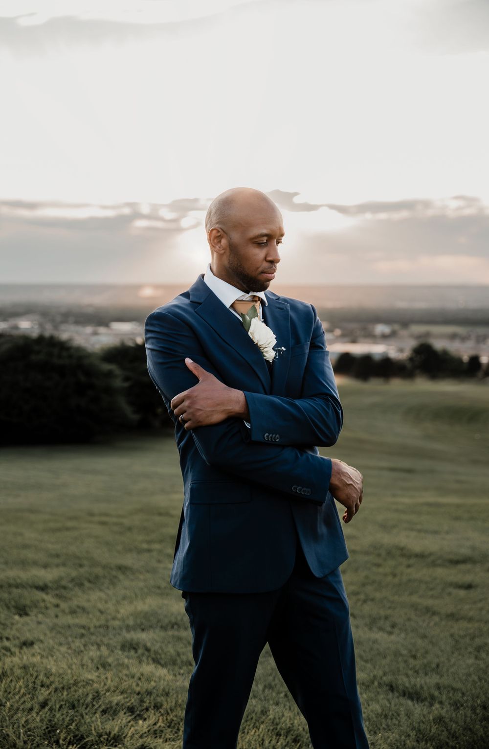 Pensive groom in blue suit stands with crossed arms at sunset on grassy hillside.