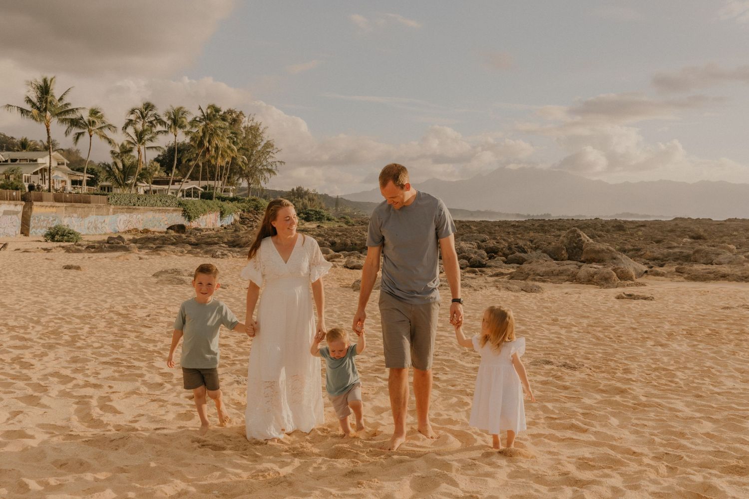 Family walking together on a sandy beach at sunset with palm trees and ocean views.