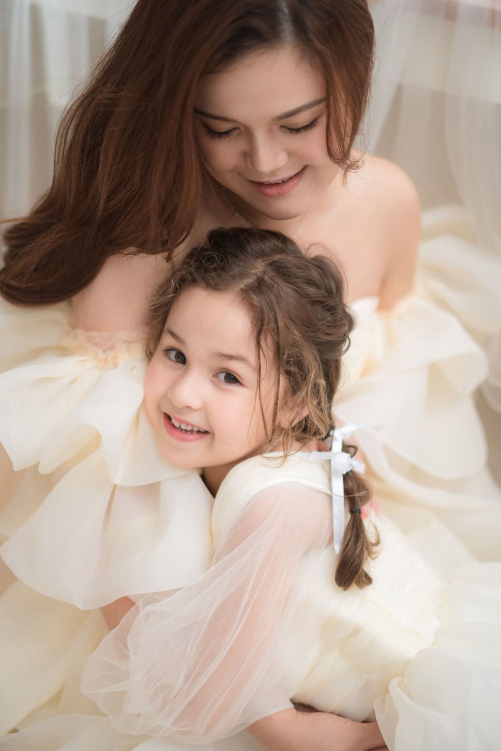 Mother and daughter in cream-colored dresses share tender moments in a bright white room.