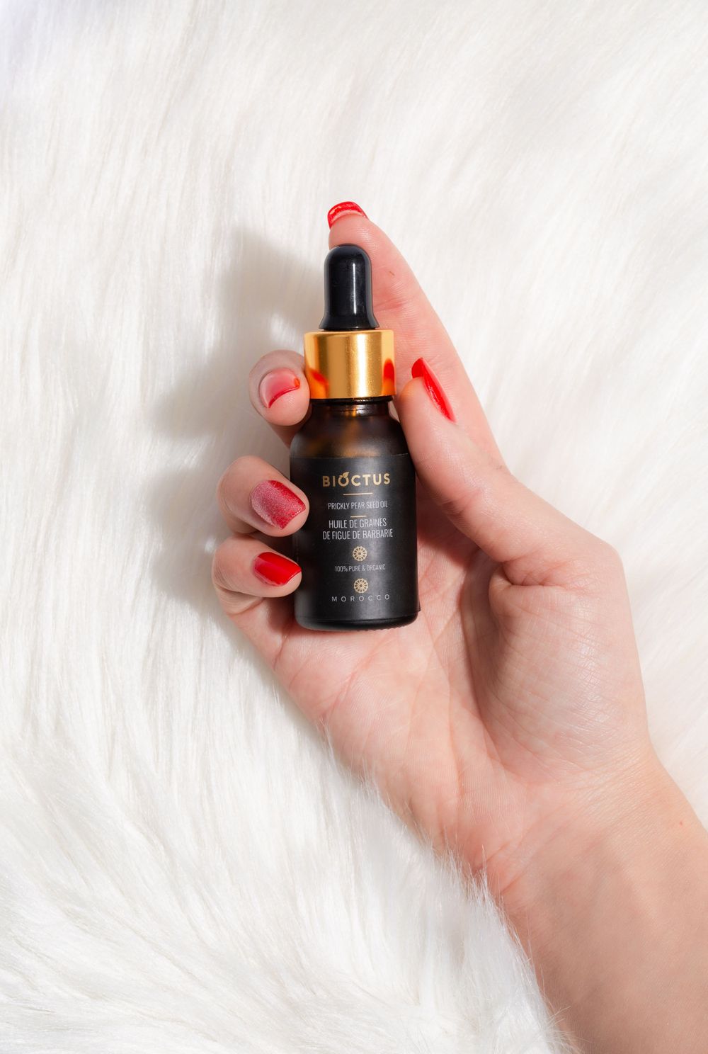 Black glass dropper bottle with red nail polish manicure on white fur background.