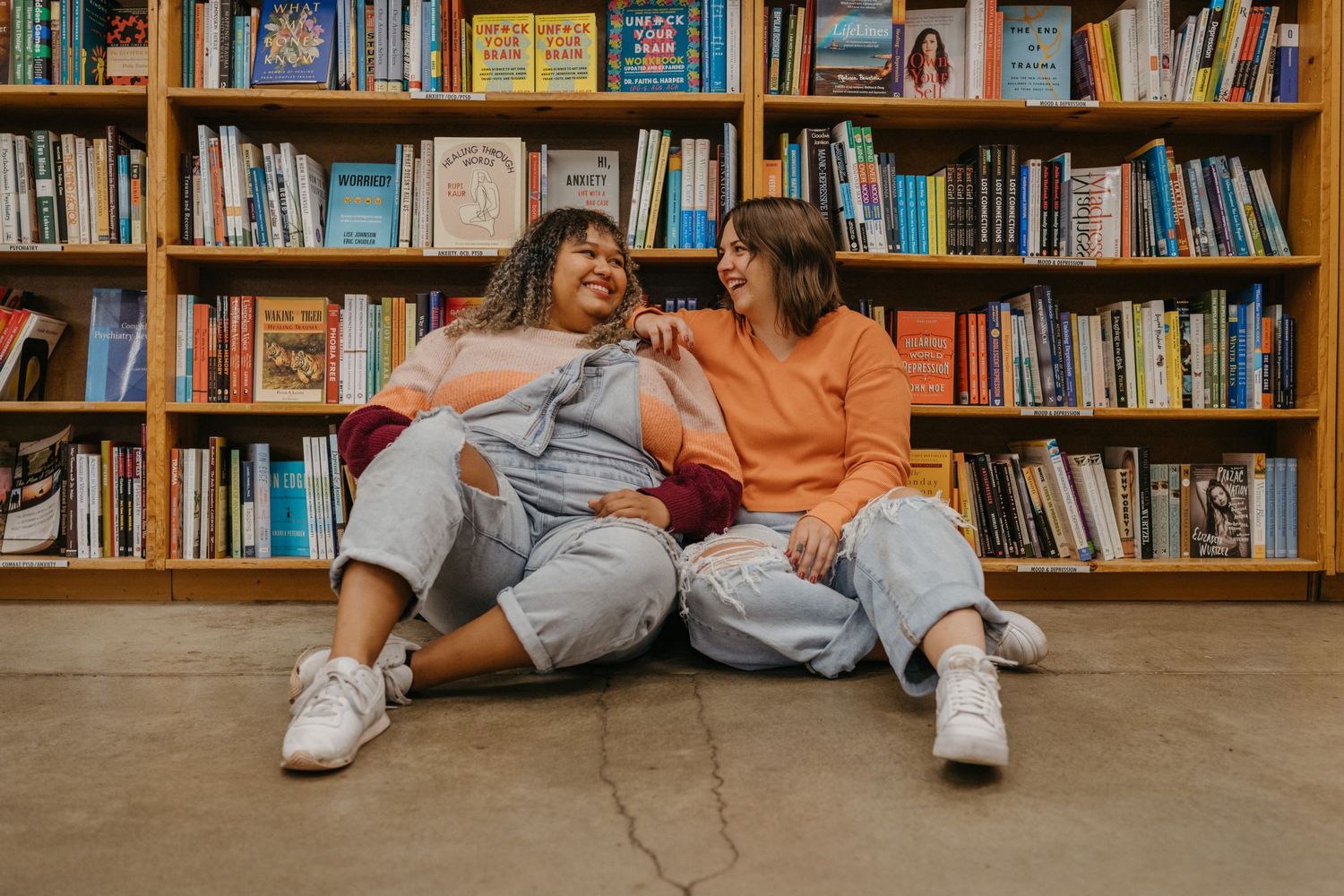Two friends laughing together while sitting on the floor of a library against wooden bookshelves.