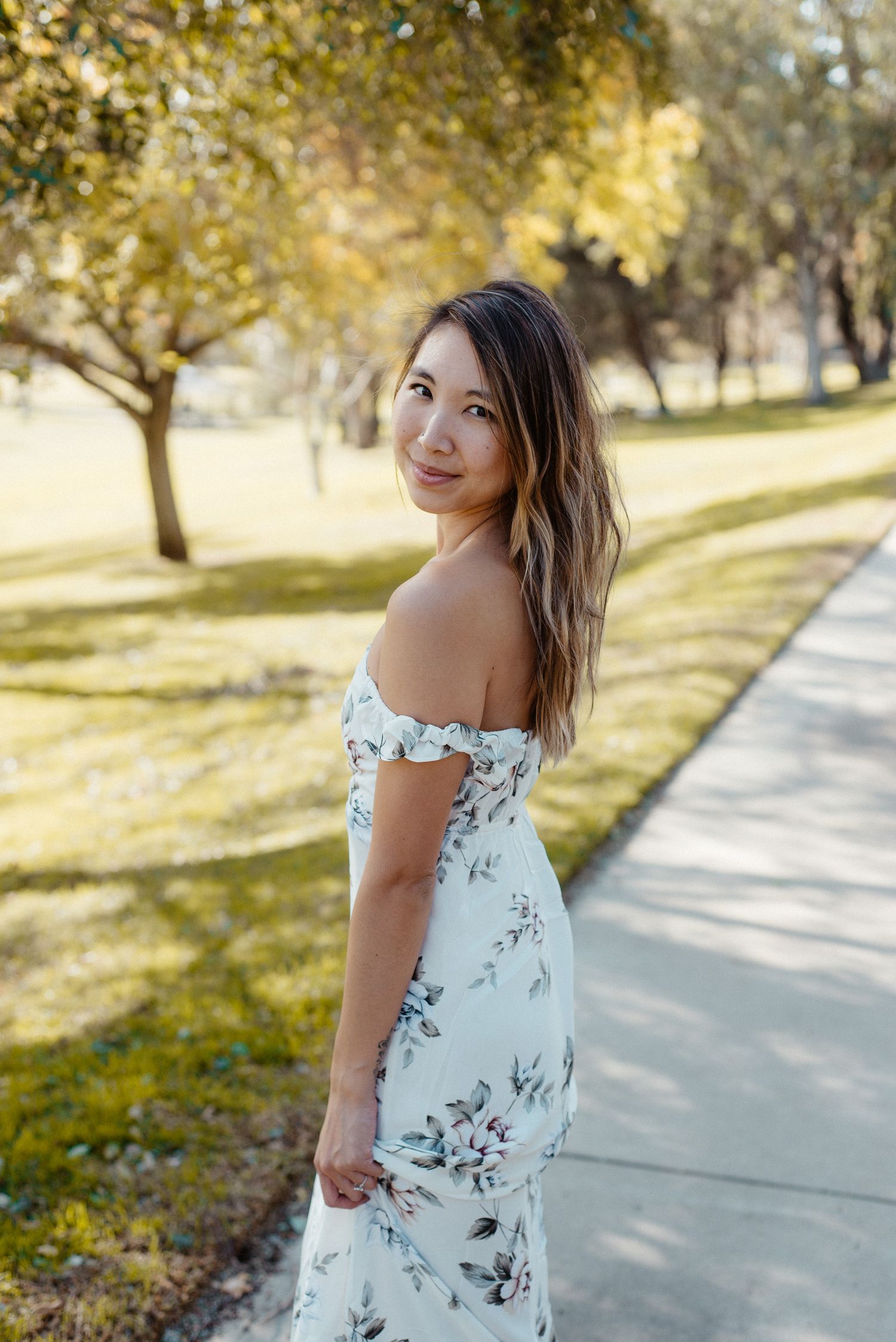 A white floral dress photographed in a sunny park setting with trees.