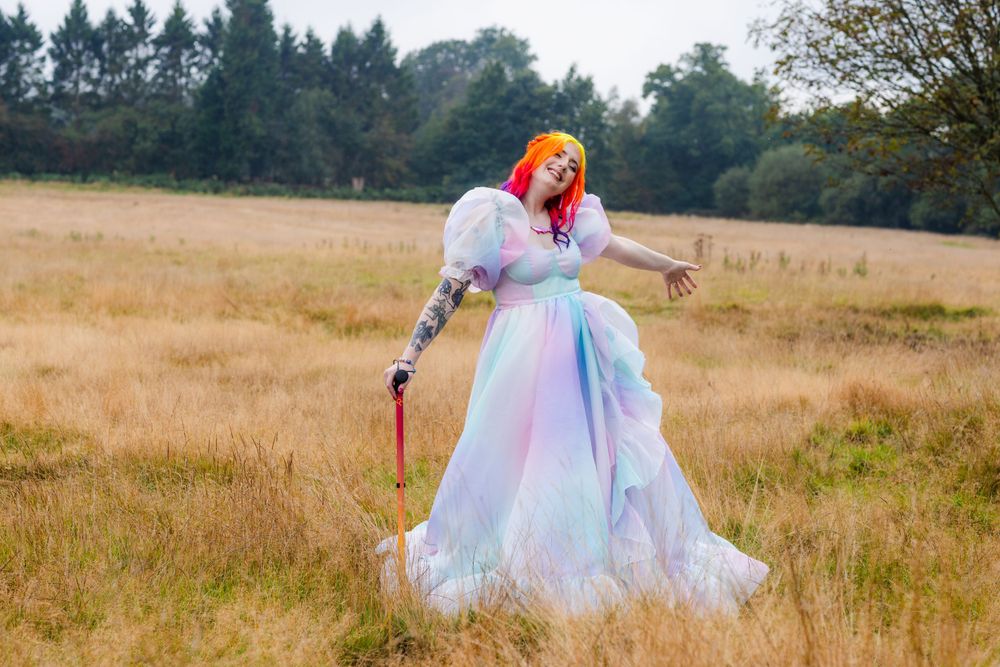 Eliza a non binary femme rainbow haired person  smiling in a field in their wedding dress with a mobility aid