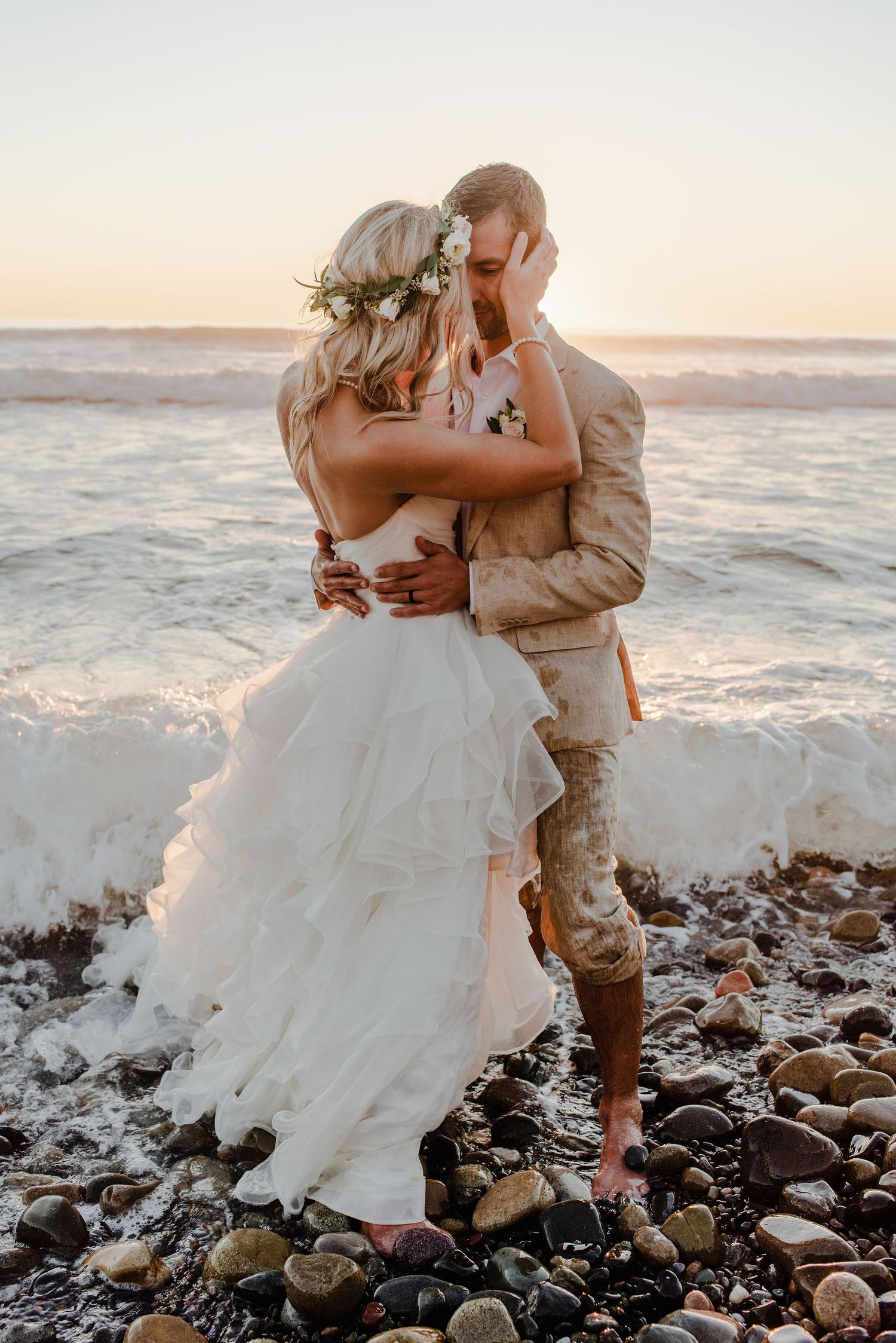 Standing in shoreline waves with wet dress and suit, bride and groom hold each other in front of the setting sun