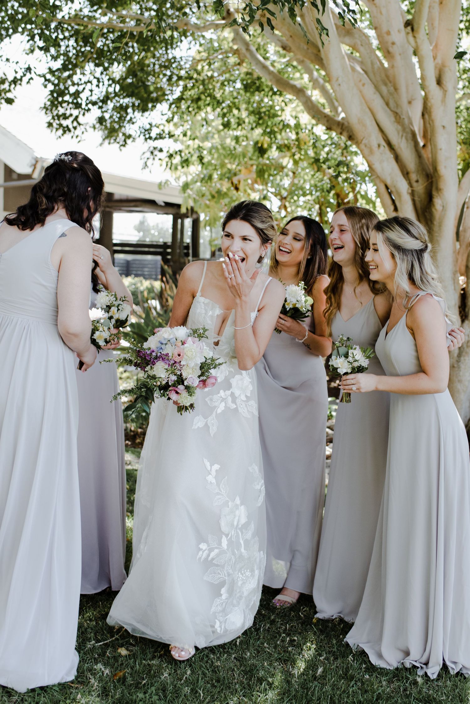 A bride holds her bouquet and looks out past her bridal party surrounding her and giggles with her hand over her mouth