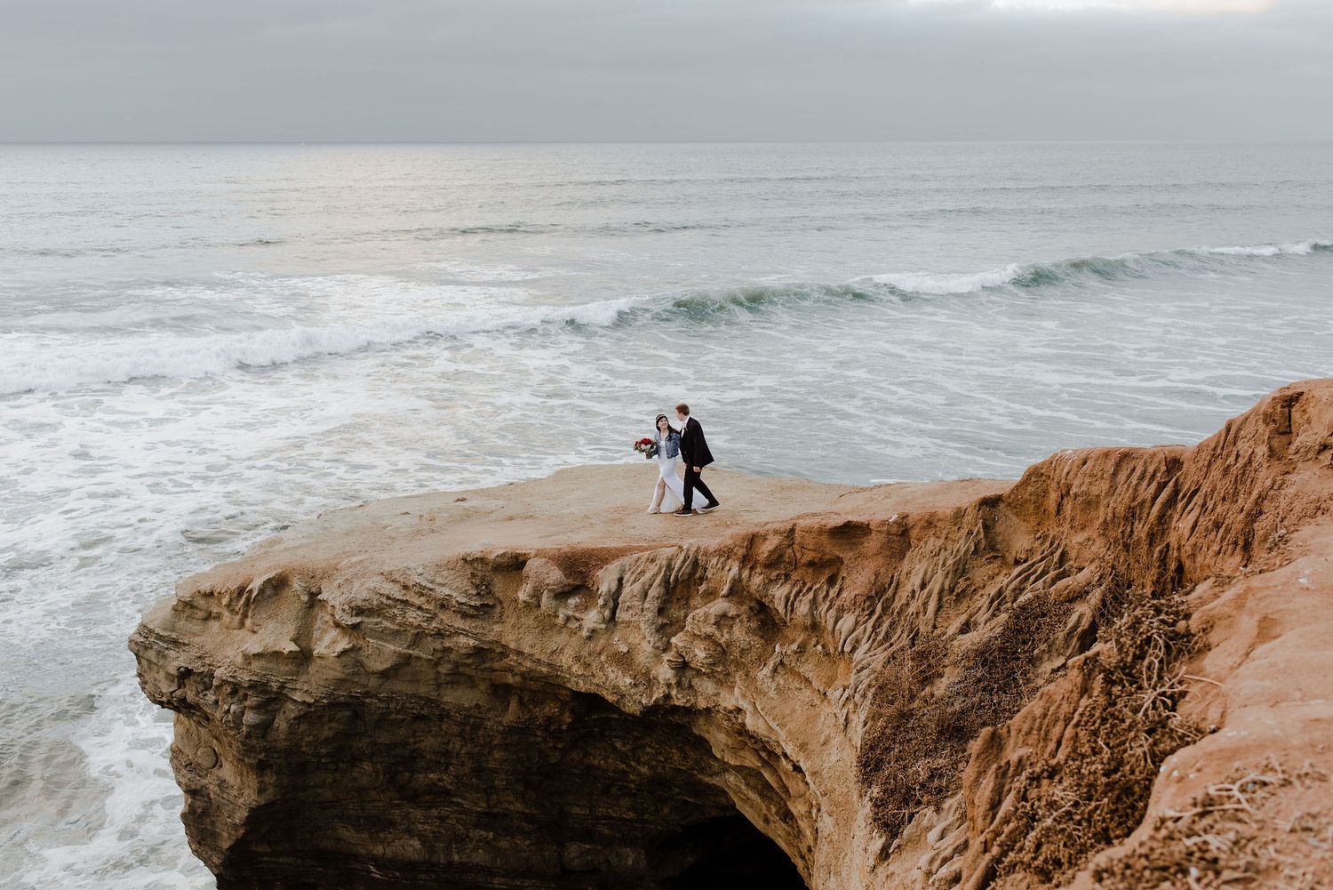 Bride and groom in the distance walk hand in hand down a clifftop overlooking the ocean