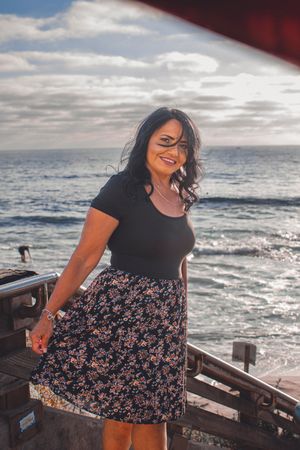 A woman in a floral dress poses on wooden stairs near the ocean during sunset.