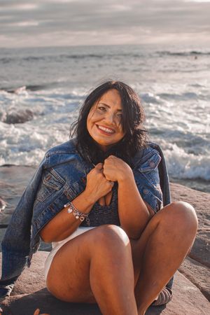 Joyful beach photoshoot featuring casual denim jacket and white shorts against ocean waves and rocky backdrop at sunset.