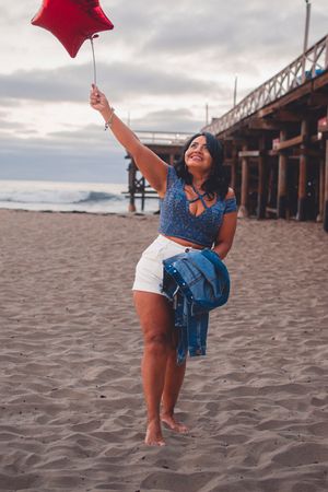 Playful beach poses with red balloon against pier backdrop during sunset photoshoot.