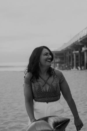 A joyful beachside portrait with someone sitting in the sand near a wooden pier as waves crash behind them.