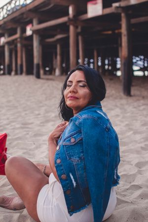 Someone relaxing on sandy beach wearing denim jacket and white shorts under a pier structure.