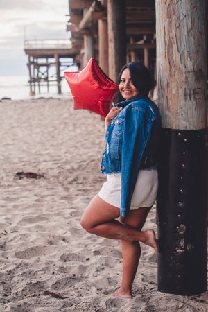 Person holding red star balloon poses playfully against wooden pier post on beach.