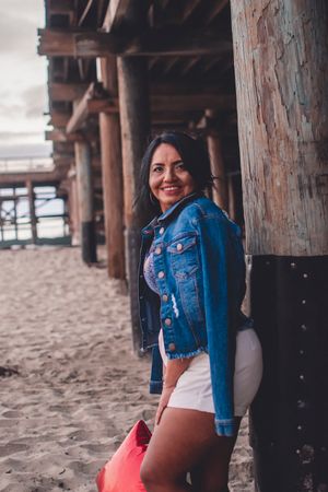 Casual beachside pose in denim jacket and white shorts against wooden pier pillars.