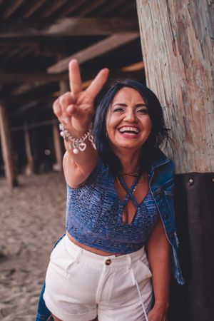 Energetic peace sign pose in blue crop top and denim jacket under beach pier.