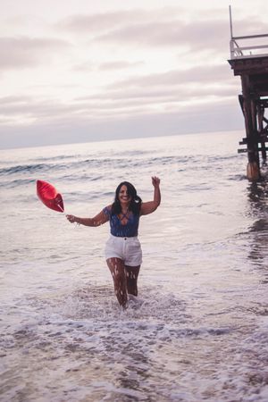 Dancing in the waves with red balloon and denim jacket near wooden pier structure.