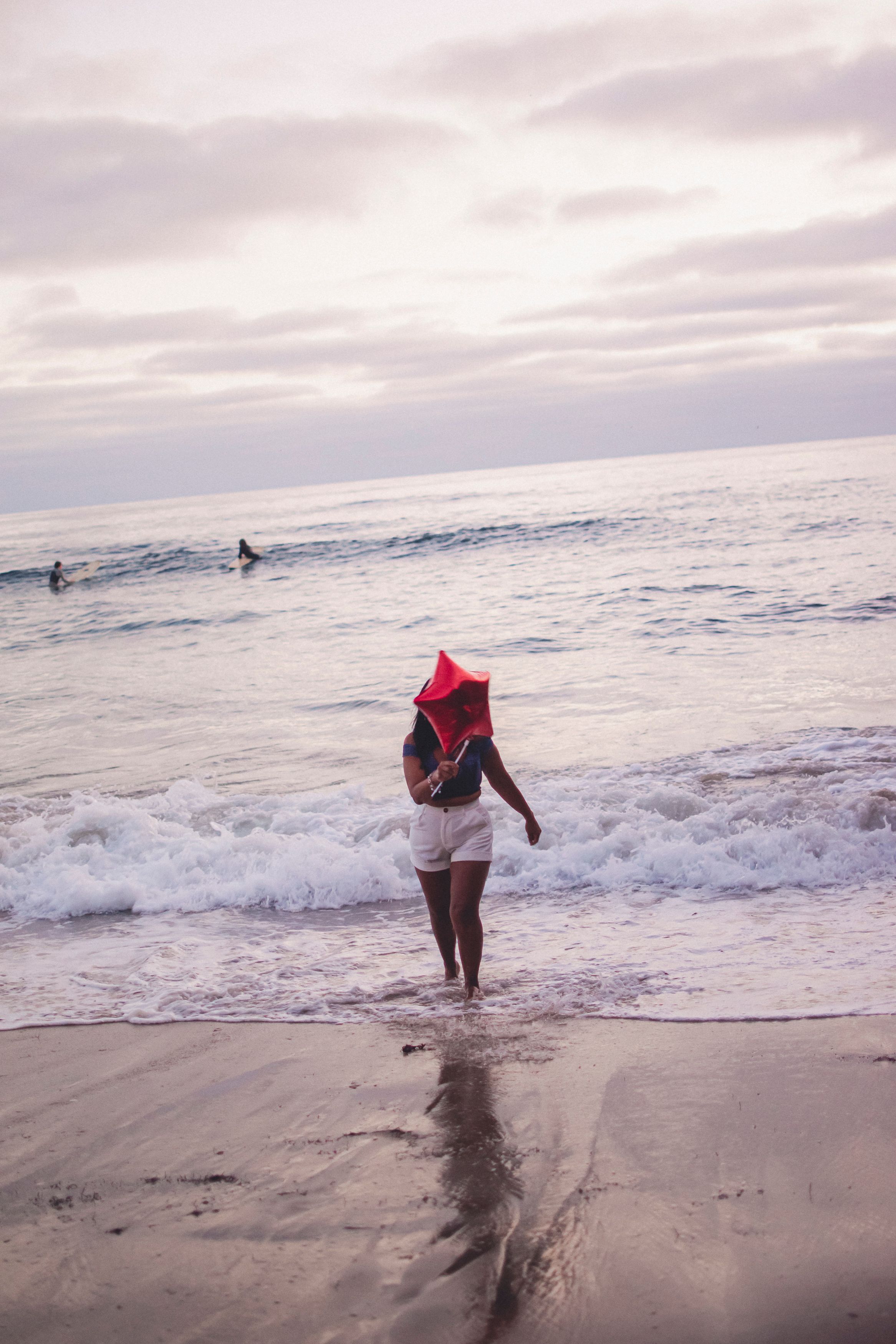 Silhouette walking into ocean waves holding red balloon with surfers in background.