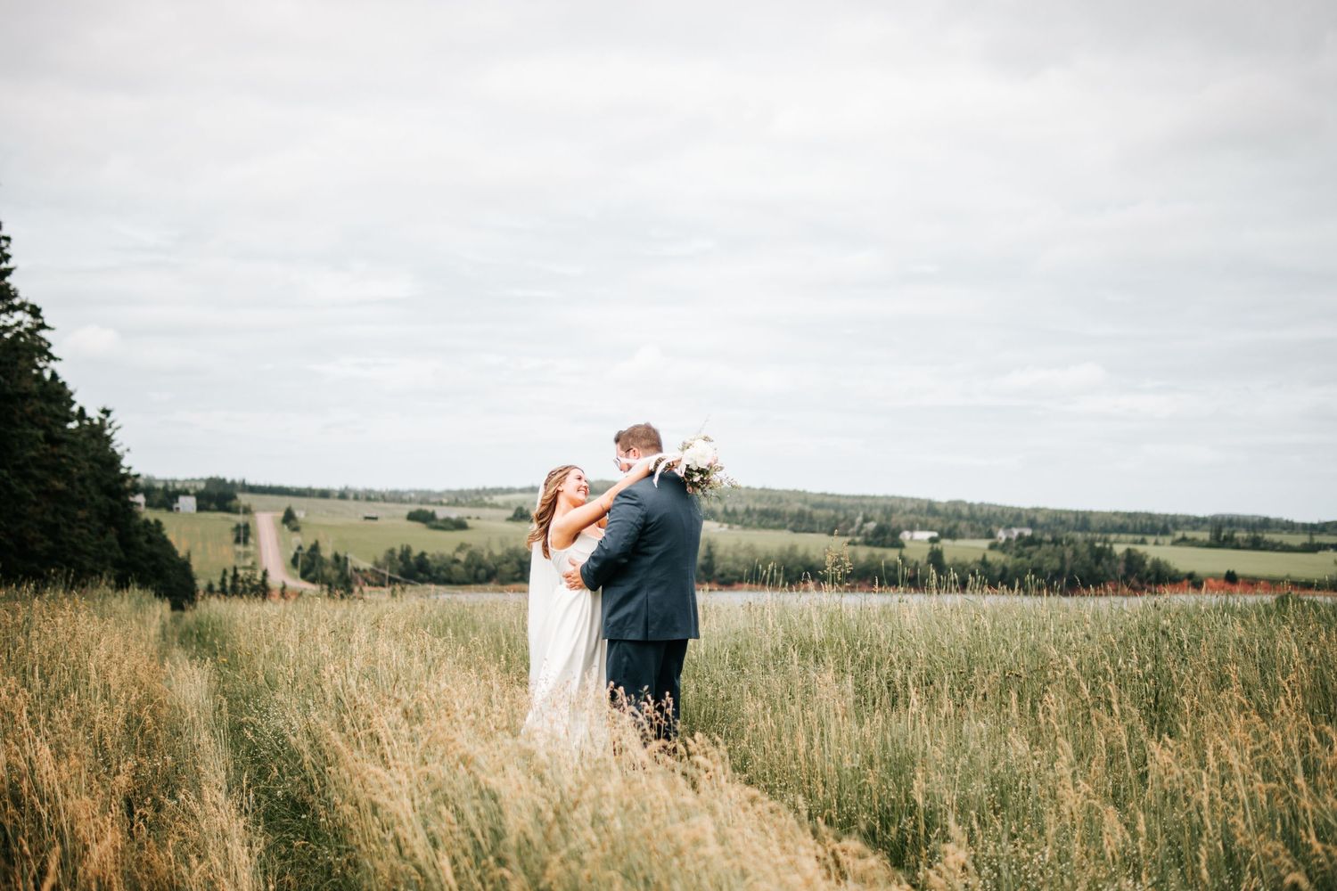 Newlywed couple embraces in a scenic meadow with rolling hills in the background. Photo at Carriage House New London PEI