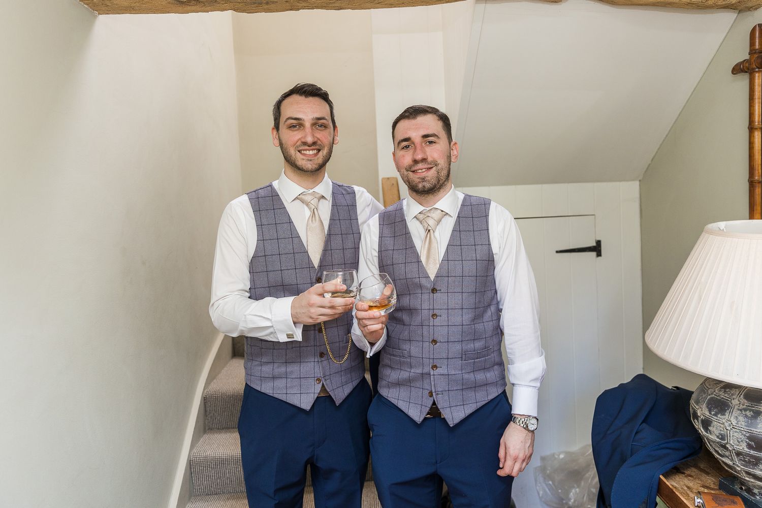 Two groomsmen in matching gray vests toast with drinks before the ceremony.