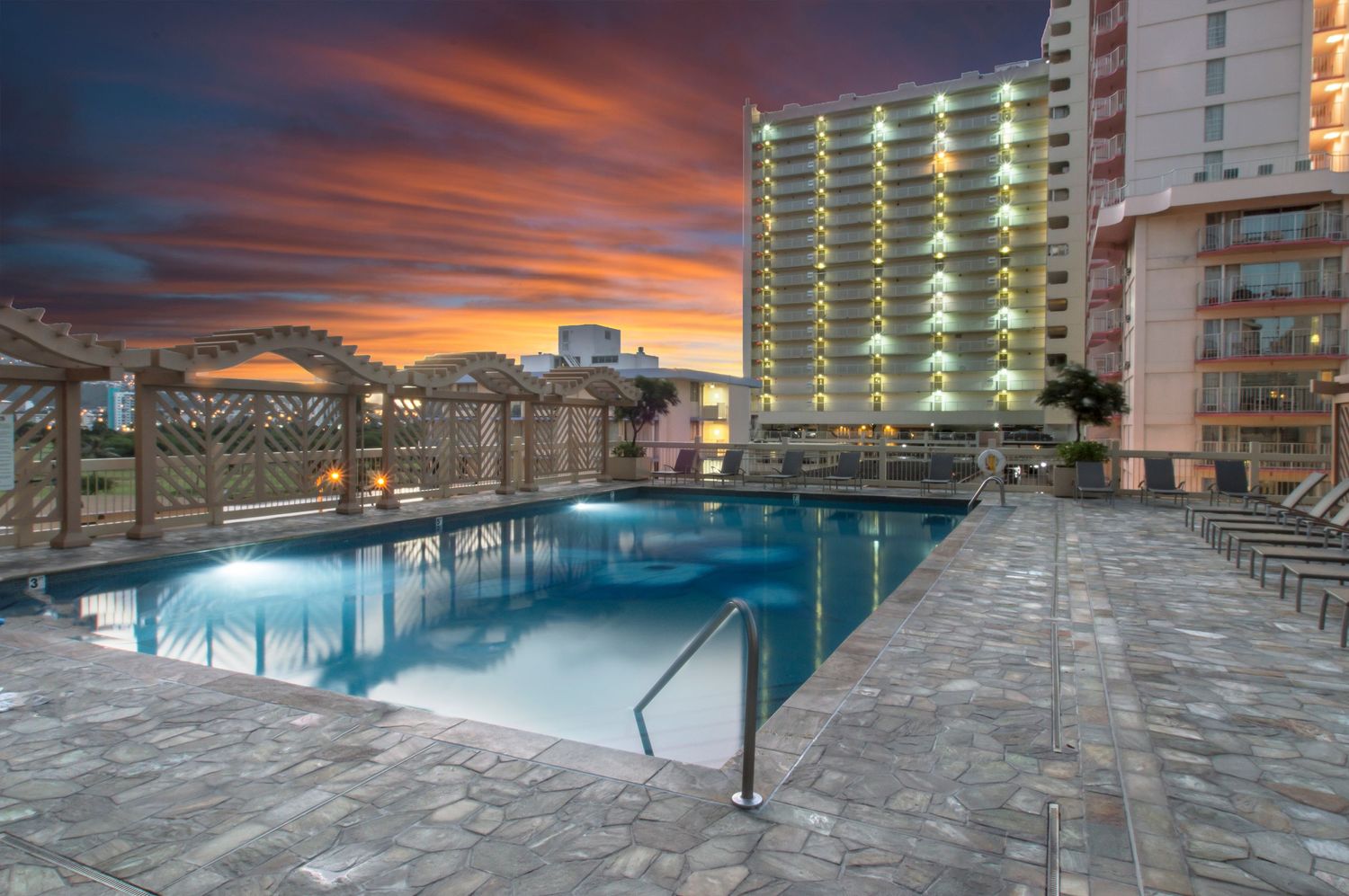 Luxurious resort pool deck with dramatic orange sunset sky and modern buildings.