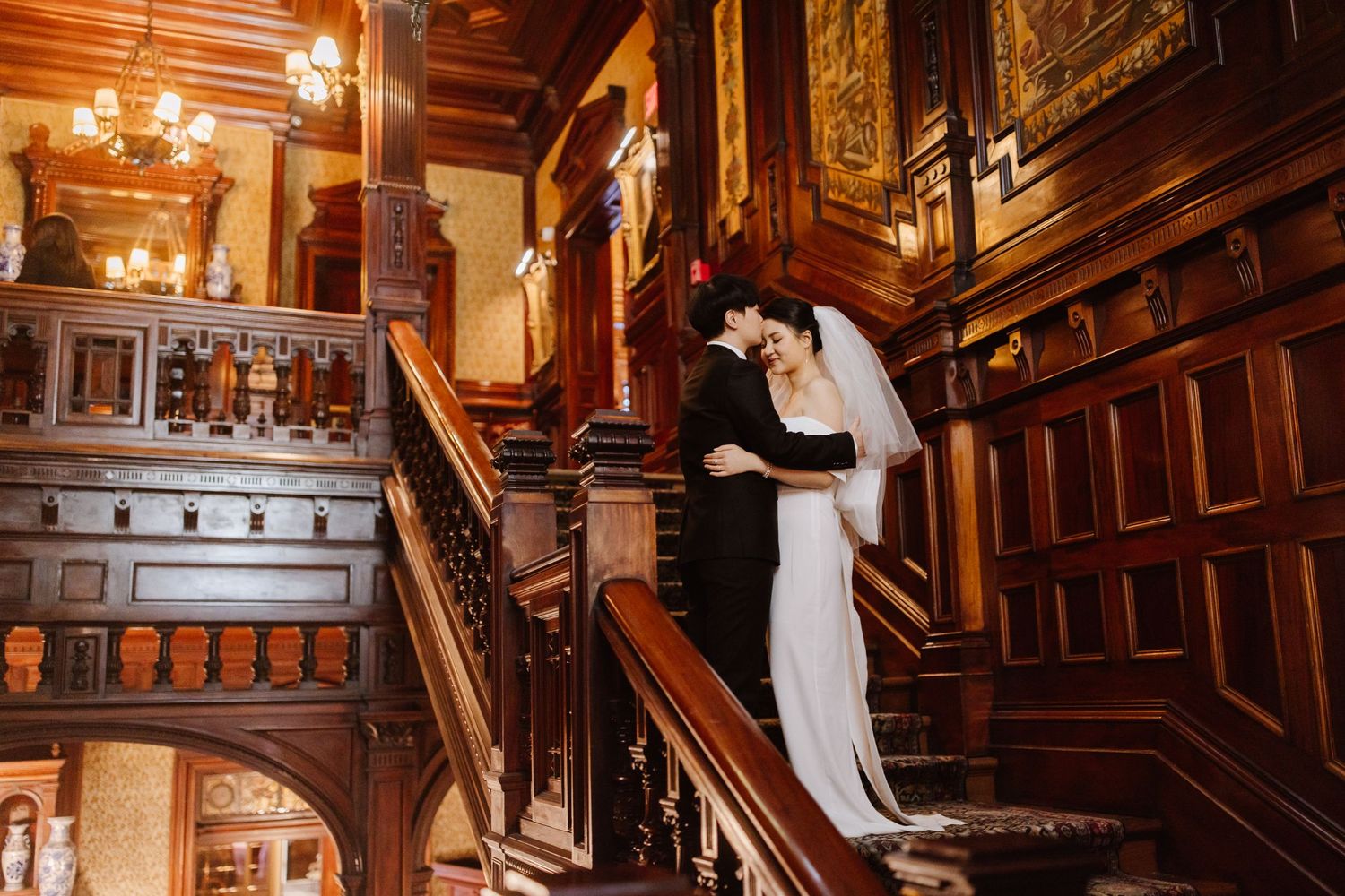 A wedding couple embraces on an ornate wooden staircase in a vintage mansion with warm lighting.