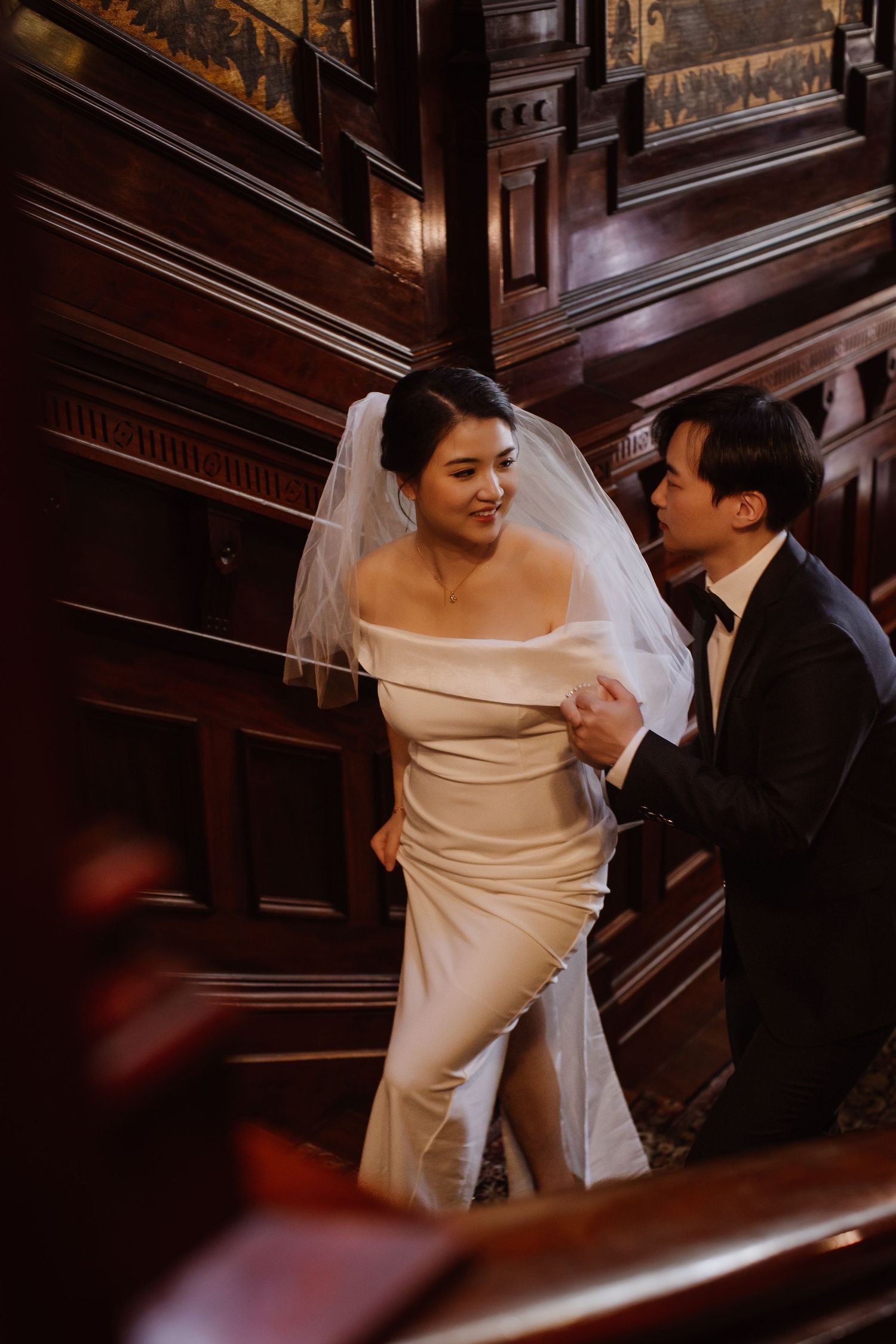 Bride in off-shoulder gown and veil shares intimate moment with groom on grand staircase.