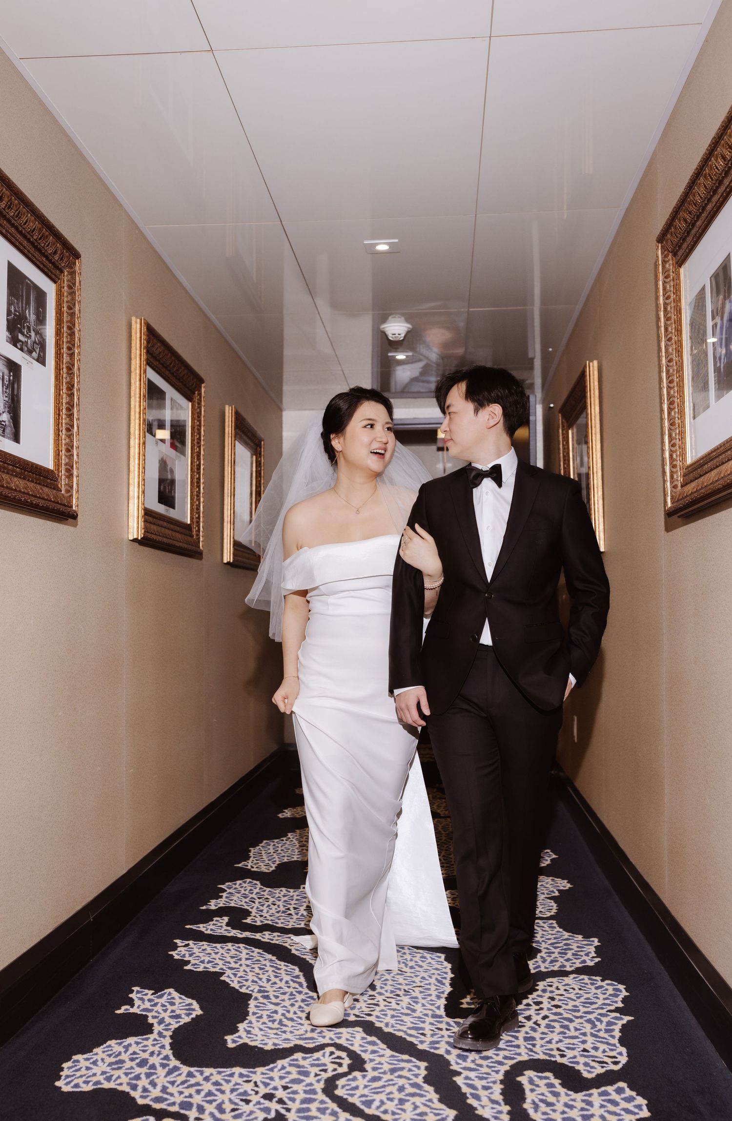 Wedding couple walks together in elegant hallway with patterned carpet and framed art.