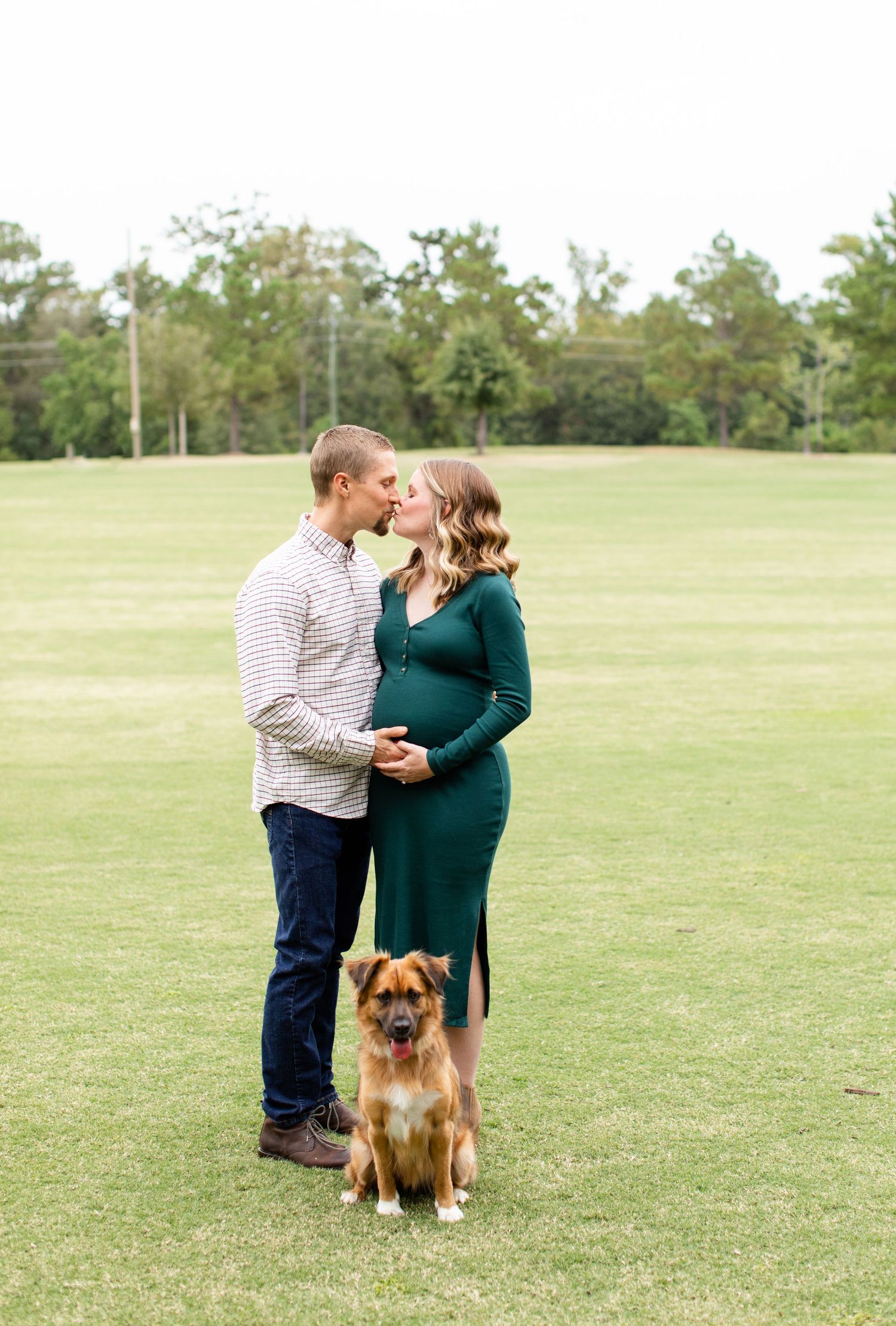 An expecting couple poses with their dog on a green lawn.