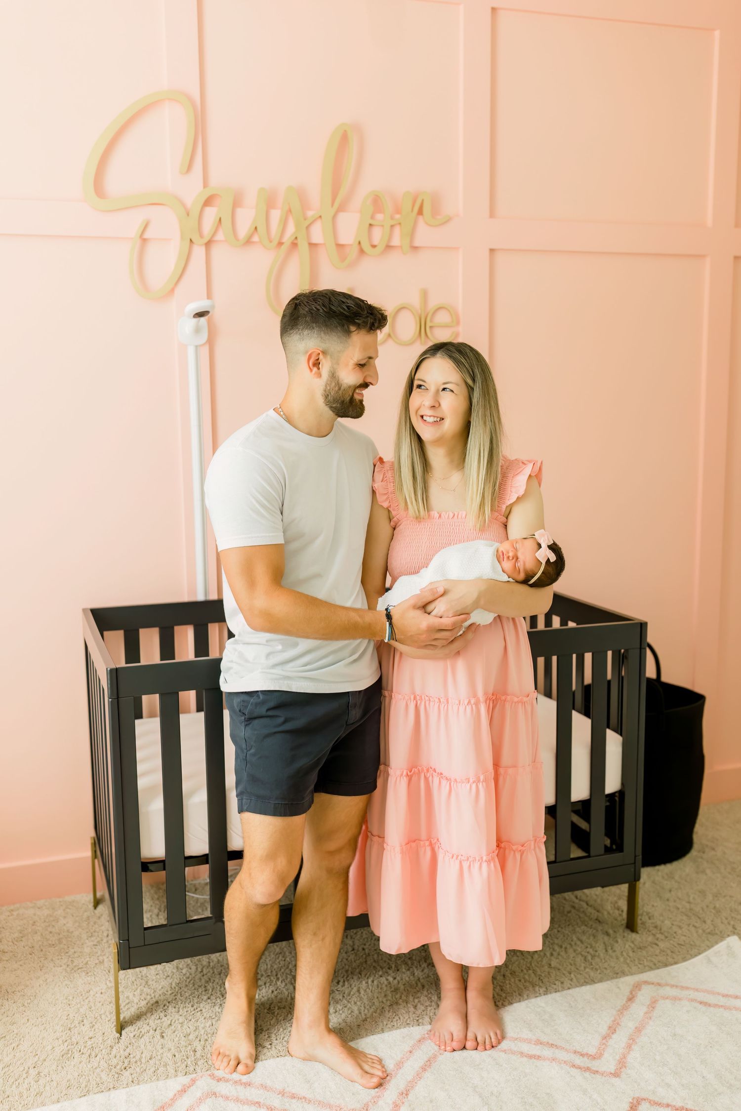 A couple holding their newborn in a nursery with pink walls and gold wall decor.