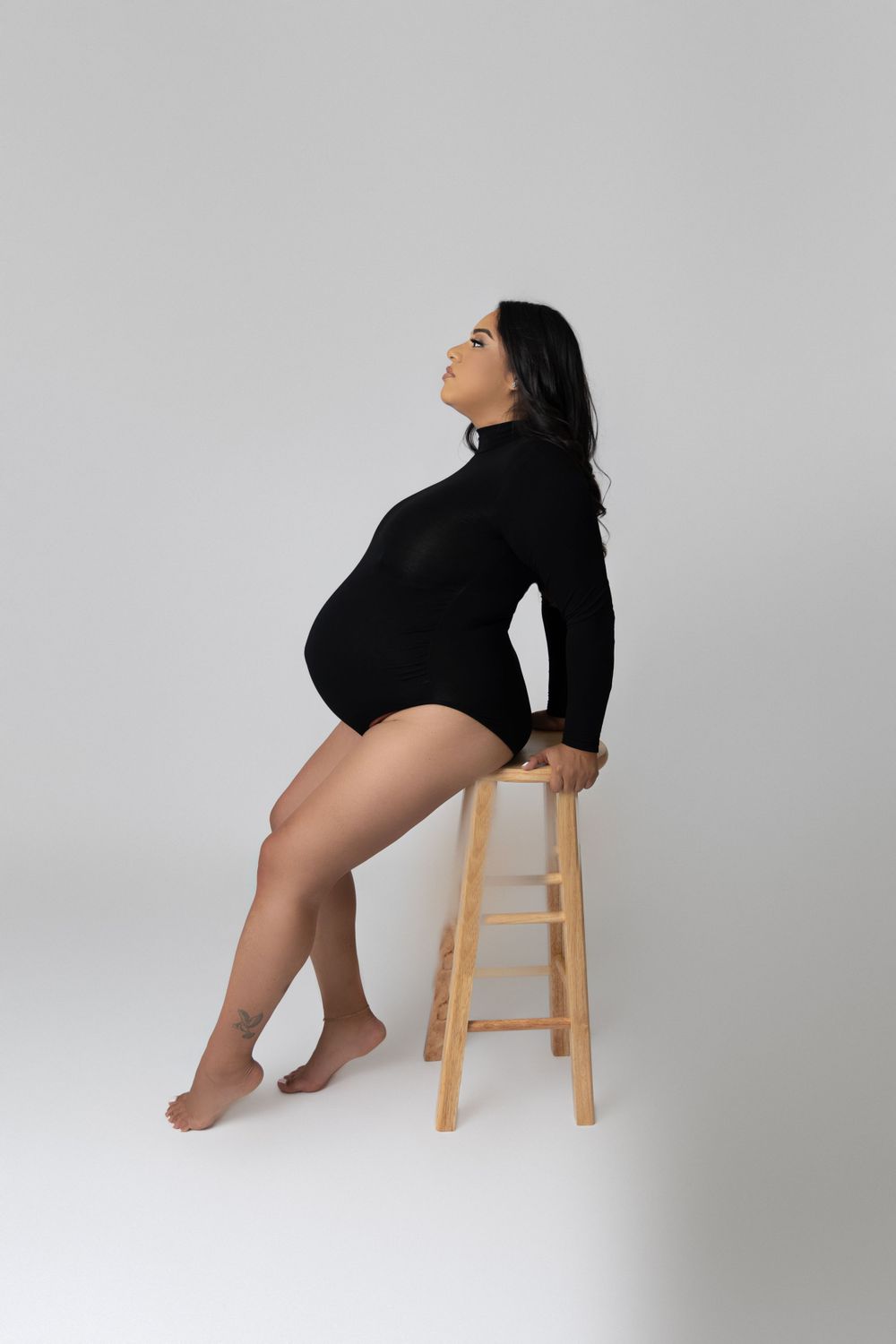 Artistic maternity portrait of woman in black bodysuit posing on wooden stool.
