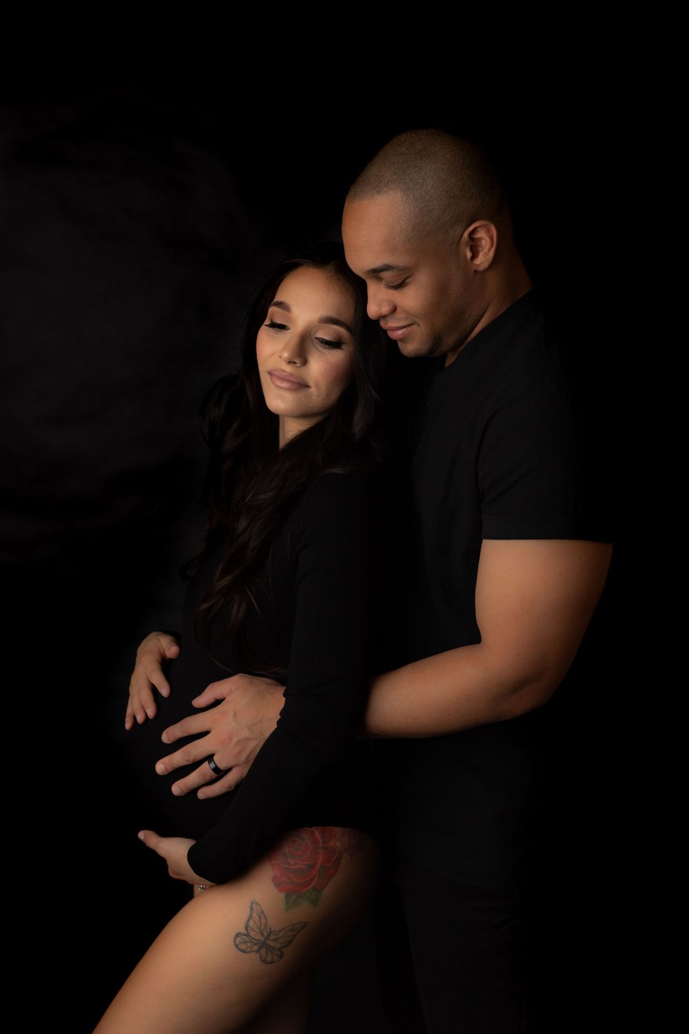 Intimate low-key maternity portrait of a couple in black clothing against a dark background.