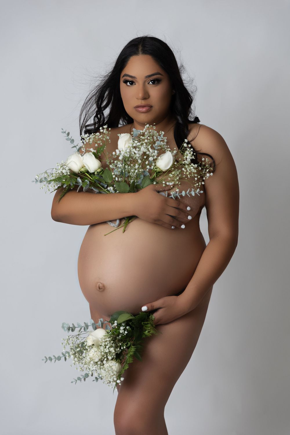 Maternity photo with white roses and baby's breath flowers against grey backdrop.