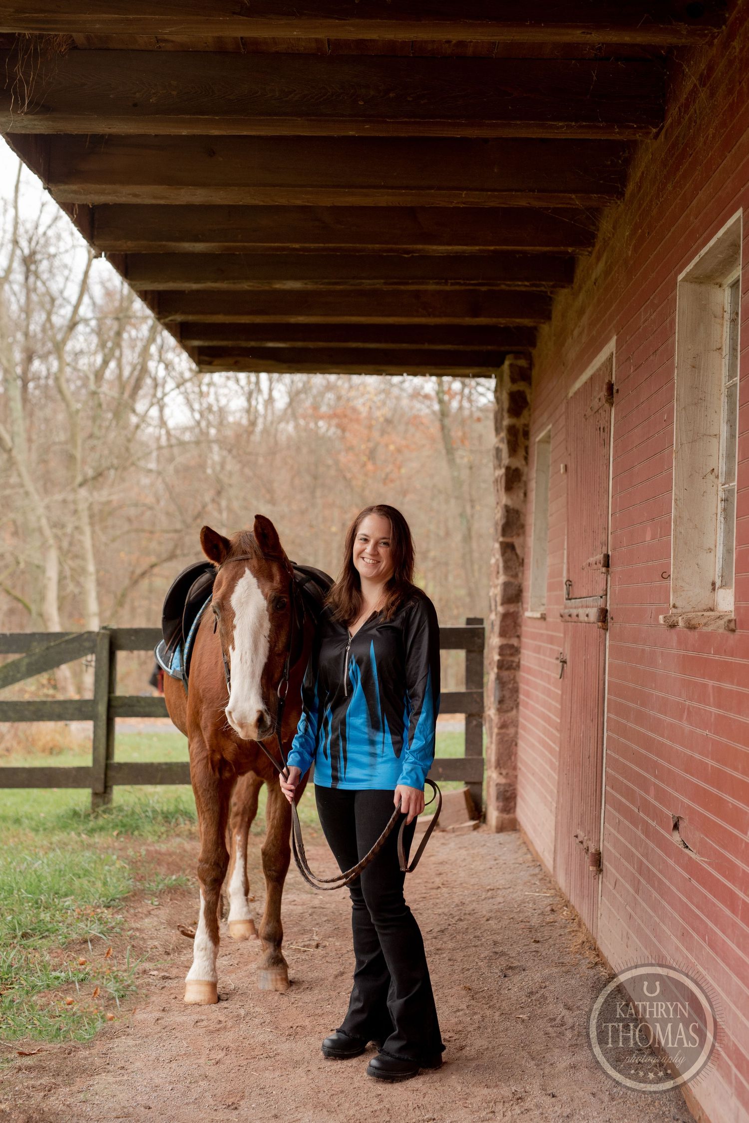 A brown horse and rider stand under a covered walkway next to a red barn building.