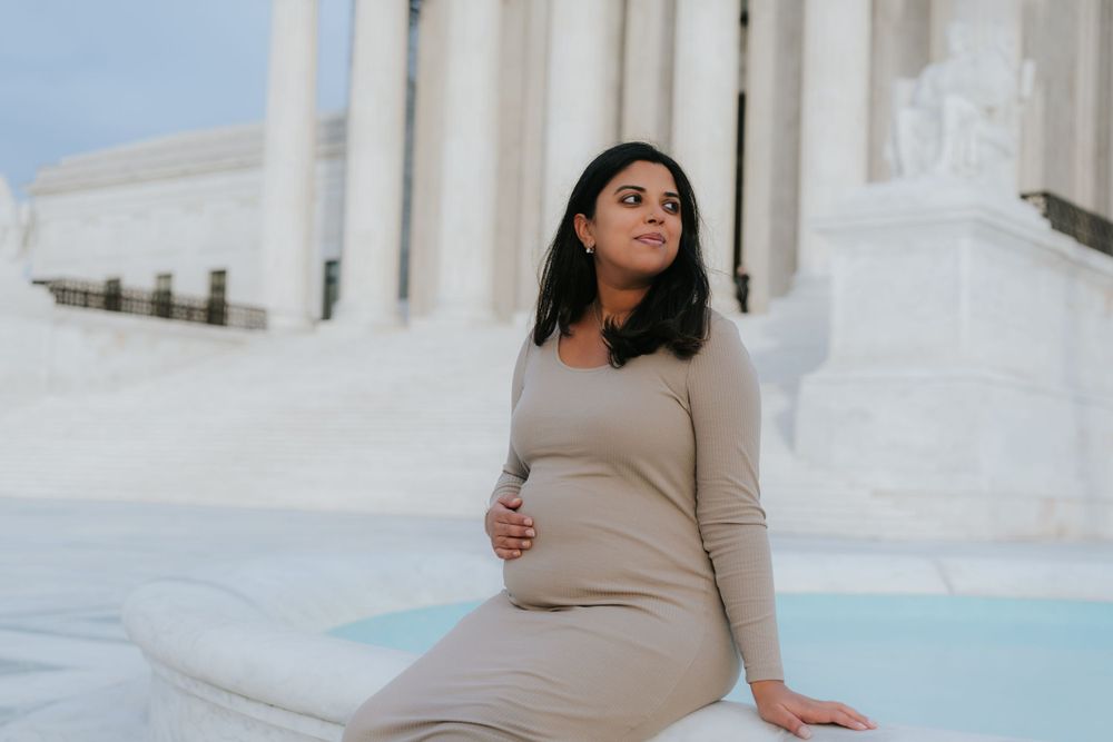 Pregnant woman in beige dress sitting by white architectural columns.