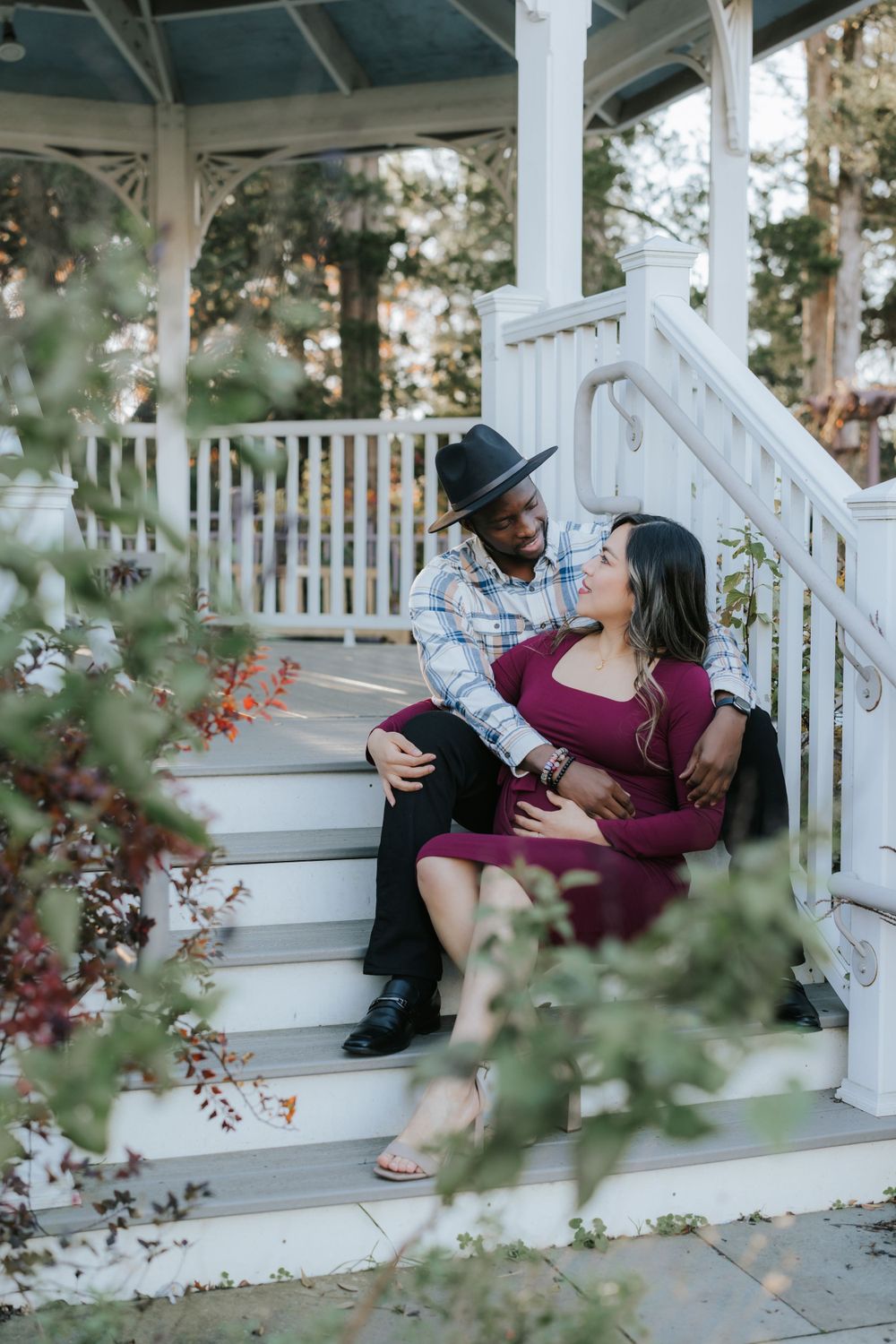 Couple shares intimate moment on white gazebo steps surrounded by flowering bushes.
