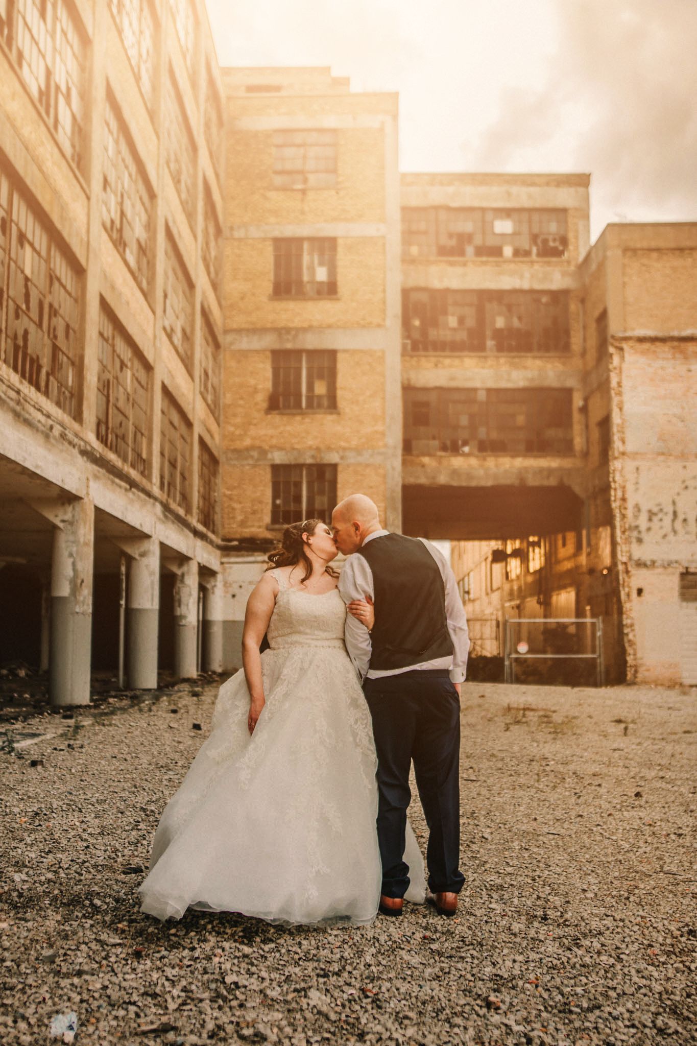 A couple in wedding attire shares an intimate moment outside an urban industrial building at sunset.