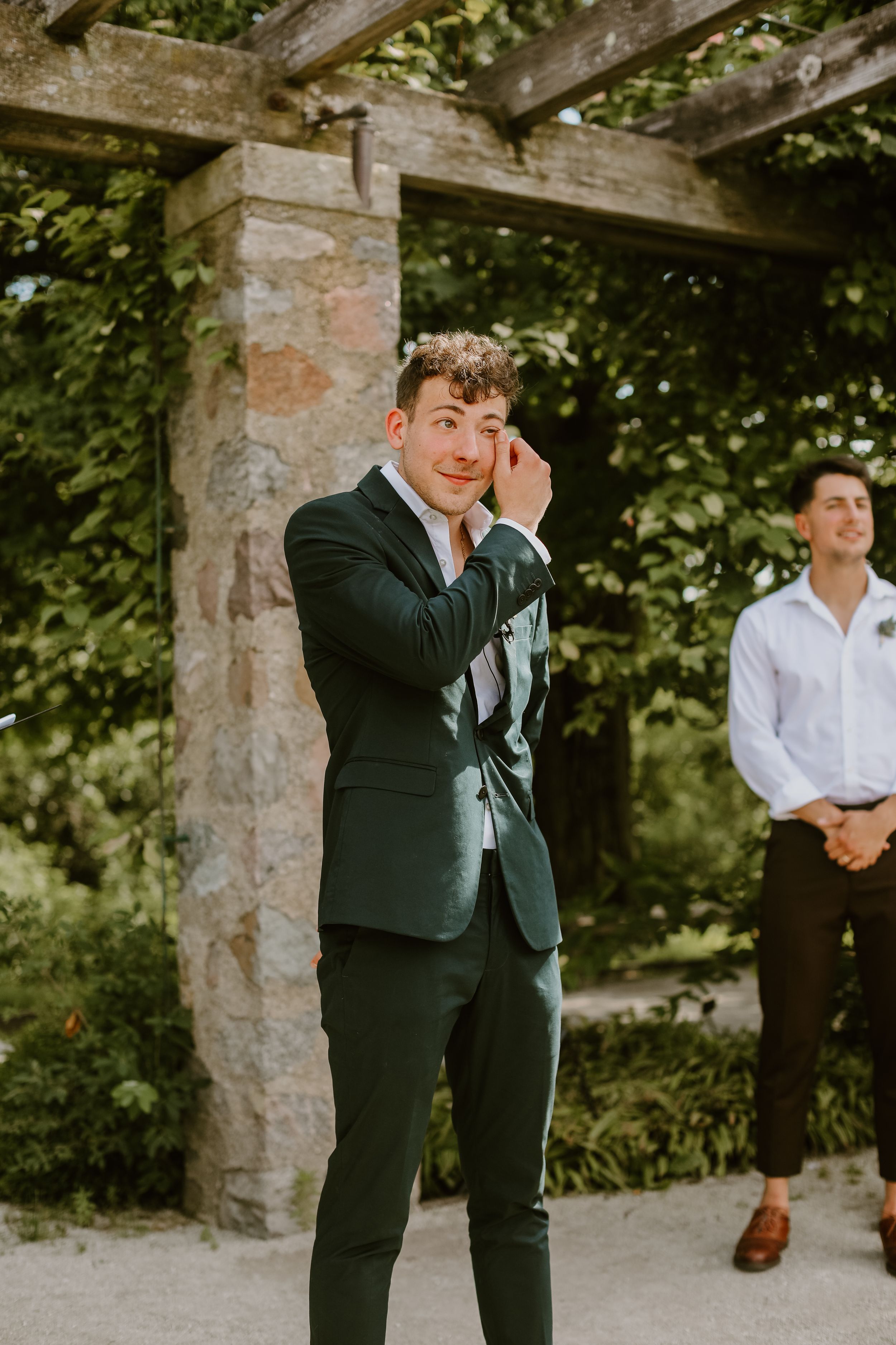A suited figure shows emotion during an outdoor wedding ceremony under a stone pergola.